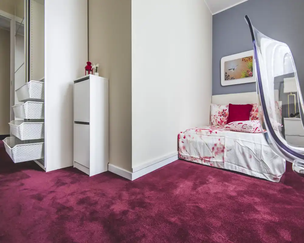 Cosy single bedroom corner with floral bedding, plush burgundy carpet and a modern transparent chair. The composition highlights the bed and bedside area, ideal for a comfortable short-stay rental.