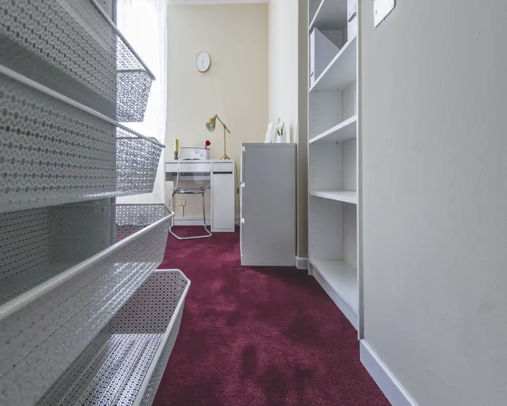 Narrow room with a small desk and shelving, carpeted floor and natural light — suitable as a home office or study corner.