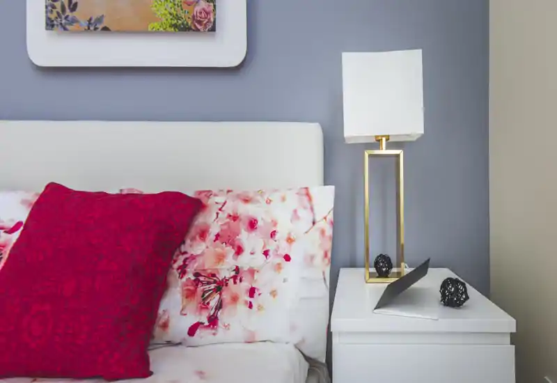 Bright, styled bedroom corner with floral bedding and decorative red cushion beside a white bedside table and modern lamp.