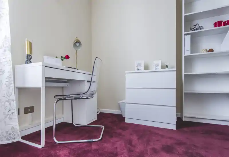 Neat home study corner with a white desk, transparent chair, dresser and shelving on a burgundy carpet; bright, sharp image showcasing a compact workspace.