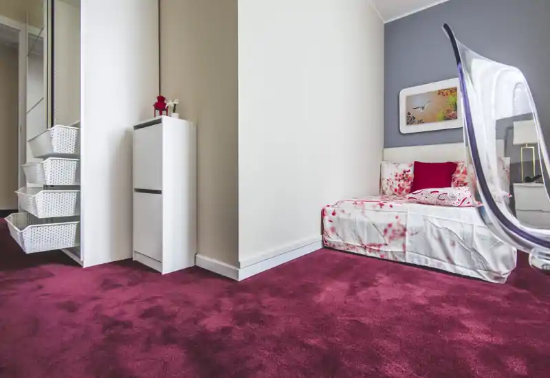 Cosy single bedroom corner with floral bedding, plush burgundy carpet and a modern transparent chair. The composition highlights the bed and bedside area, ideal for a comfortable short-stay rental.