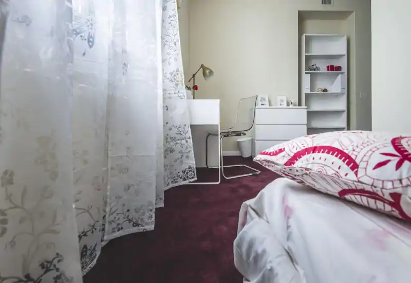 Bright, cozy bedroom corner with patterned pillows and a visible bed in the foreground, a small white desk and shelving in the background; soft natural light filters through decorative curtains.