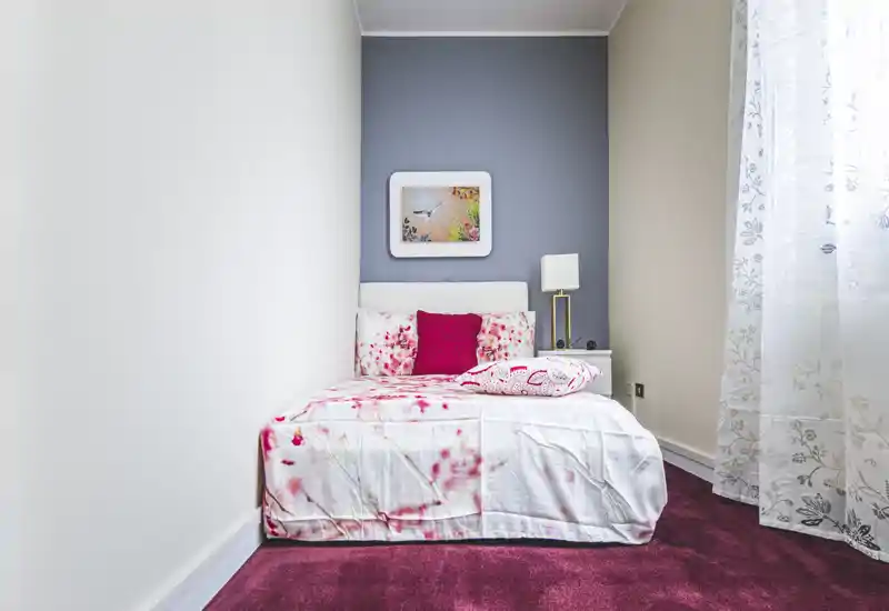 Compact, well-styled single bedroom with a neatly made bed, decorative cushions and soft natural light from a sheer-curtained window.