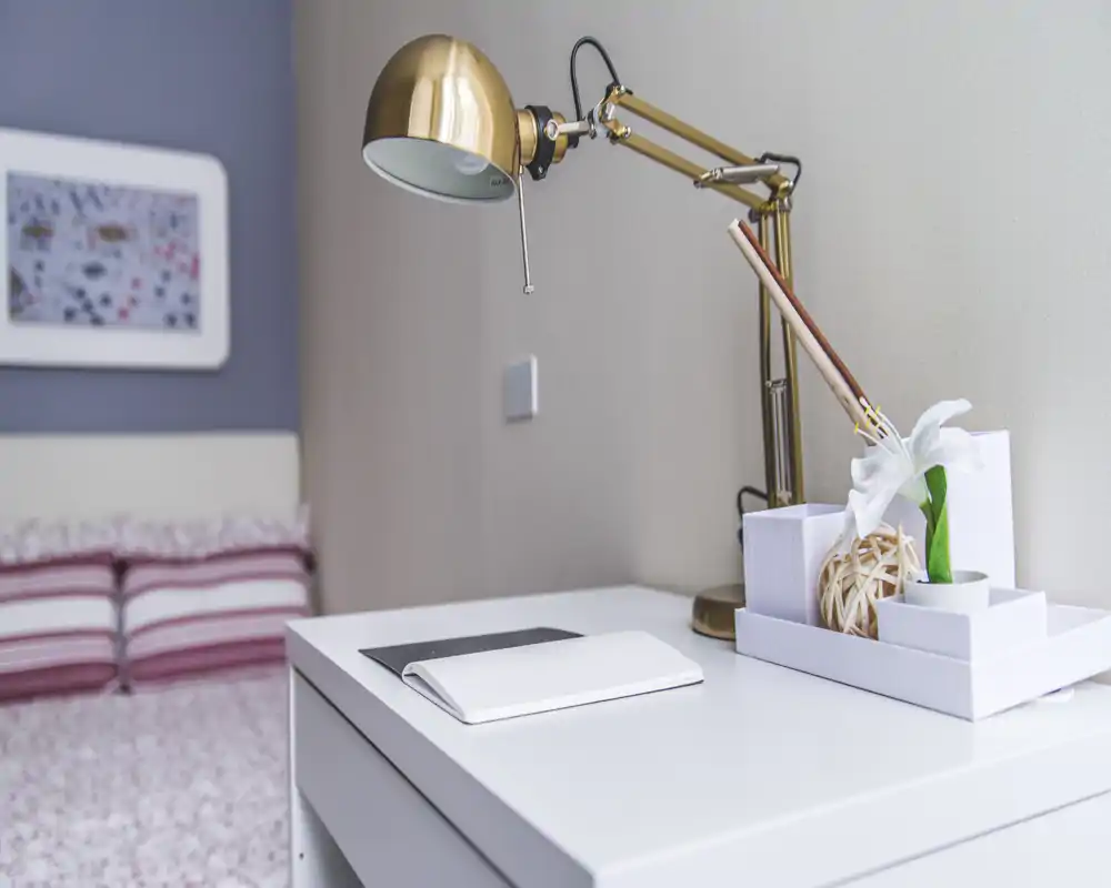 Dettaglio di una camera luminosa e moderna: scrivania da comodino con lampada dorata, vassoio decorativo e letto parzialmente visibile con cuscini a righe — un angolo pulito e accogliente ideale per lavorare o leggere.