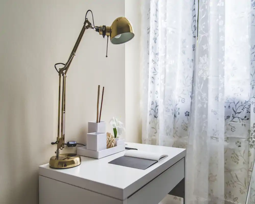 Angolo camera luminoso e minimal con scrivania bianca, dettagli decorativi e una lampada regolabile in ottone accanto a una tenda leggera con pattern.