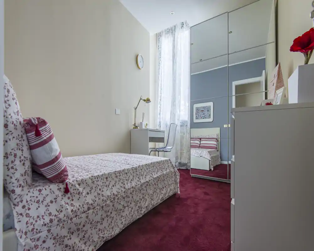 Camera singola luminosa e accogliente con biancheria floreale, una scrivania vicino a una finestra alta e un armadio con ante a specchio che amplifica gli spazi. Tappeto rosso e arredi curati rendono l'ambiente invitante.