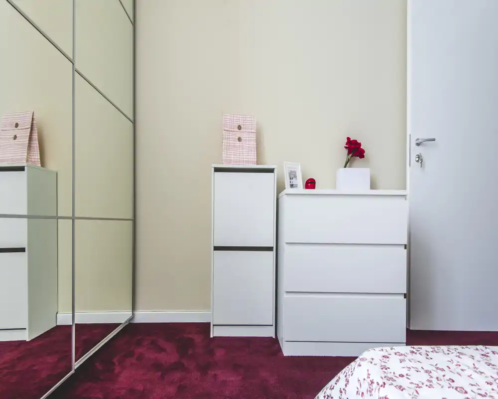 Angolo di camera da letto luminoso e minimal: comodini bianchi, ante a specchio e parte del letto con copriletto a fantasia in primo piano; moquette bordeaux e un piccolo vaso decorativo.