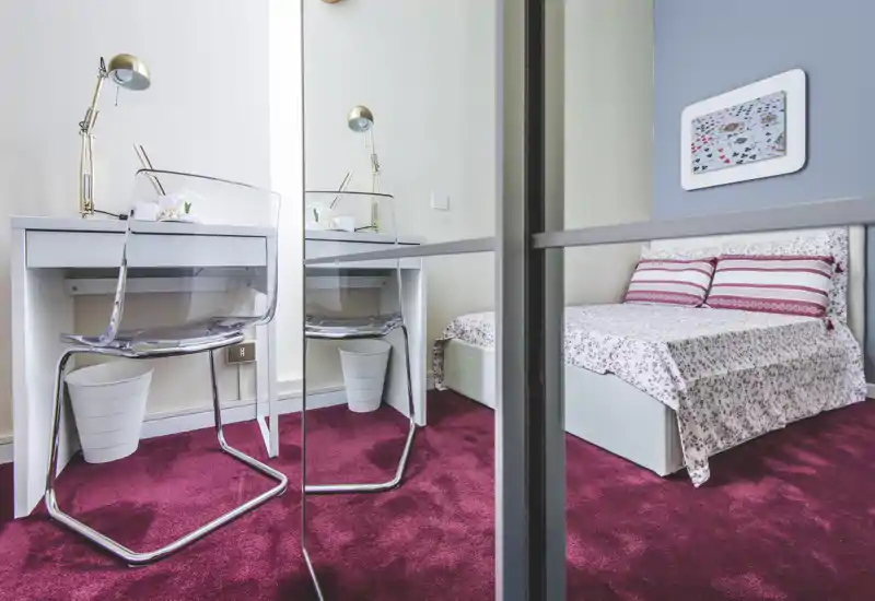 Camera da letto luminosa e ordinata con letto con copriletto a fantasia, cuscini a righe e una piccola scrivania bianca accanto a un armadio con specchio. La moquette fucsia e la biancheria creano un ambiente caldo e di stile.
