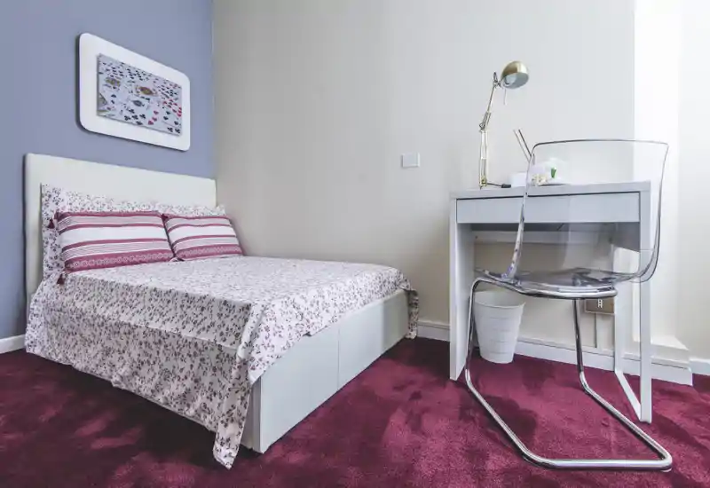 Camera da letto luminosa e ordinata con letto matrimoniale in biancheria fantasia, una piccola scrivania con sedia trasparente e lampada da lettura — ideale per una o due persone.