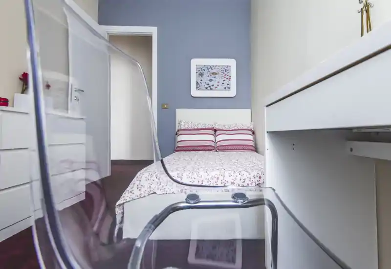 Camera da letto compatta e luminosa con letto matrimoniale in copriletto a fantasia e cuscini rigati; una sedia acrilica trasparente e una scrivania bianca creano un ambiente moderno e ordinato.
