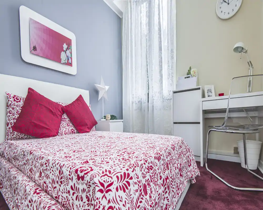 Dormitorio individual luminoso y acogedor con ropa de cama roja y blanca estampada, cojines decorativos y un pequeño escritorio junto a la ventana — ideal para una estancia agradable.
