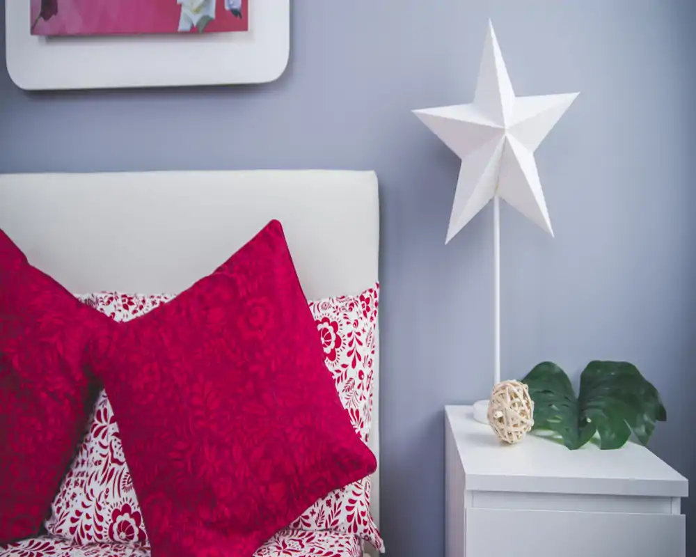 Rincón de dormitorio nítido y estilizado con cabecero blanco, ropa de cama estampada y cojines rojos vibrantes junto a una mesita con lámpara decorativa y follaje.