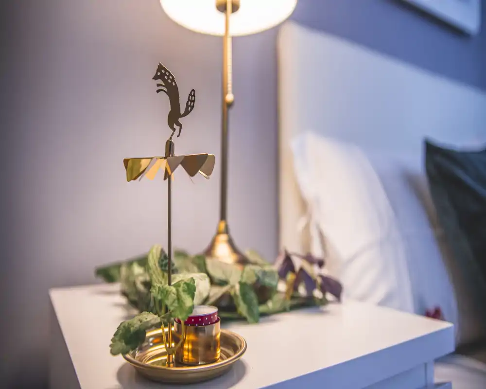 Close-up of a bedside table in a bedroom featuring a decorative lamp, small plant and candle on a crisp white nightstand; soft warm lighting and a partially visible pillow/headboard create a cozy atmosphere.