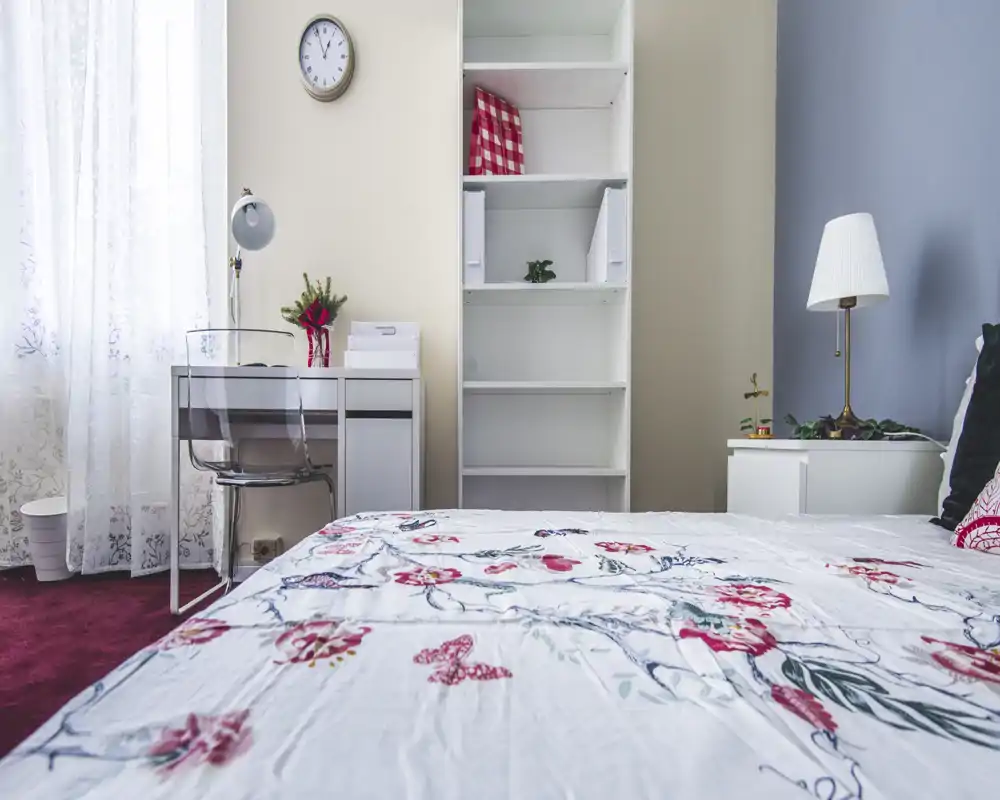 Bright, tidy bedroom with floral bedding, a bedside lamp and a small desk by the window. The bed dominates the foreground and the room has simple, modern storage and natural light.