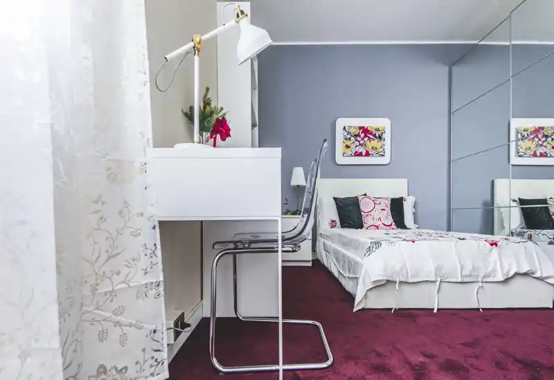 Bright, well-staged bedroom with a double bed dressed in patterned linens, decorative cushions and a bedside lamp; a desk and transparent chair occupy the foreground while mirrored wardrobe panels expand the space.