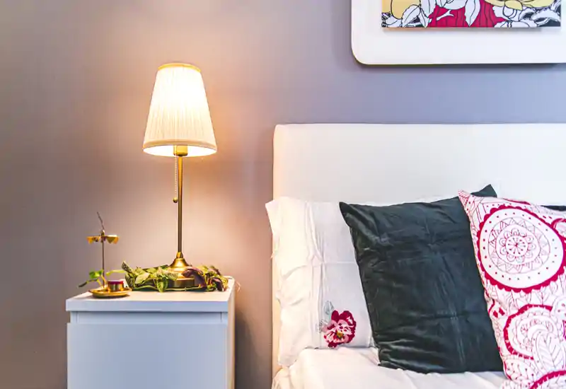 Bright, styled bedroom corner with a visible headboard, pillows and a lit bedside lamp creating a cozy atmosphere.