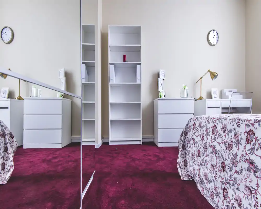 Chambre lumineuse et soignée avec couvre-lit floral, tables de nuit blanches assorties et étagère centrale ; tapis rouge profond et grand miroir apportent profondeur et style.