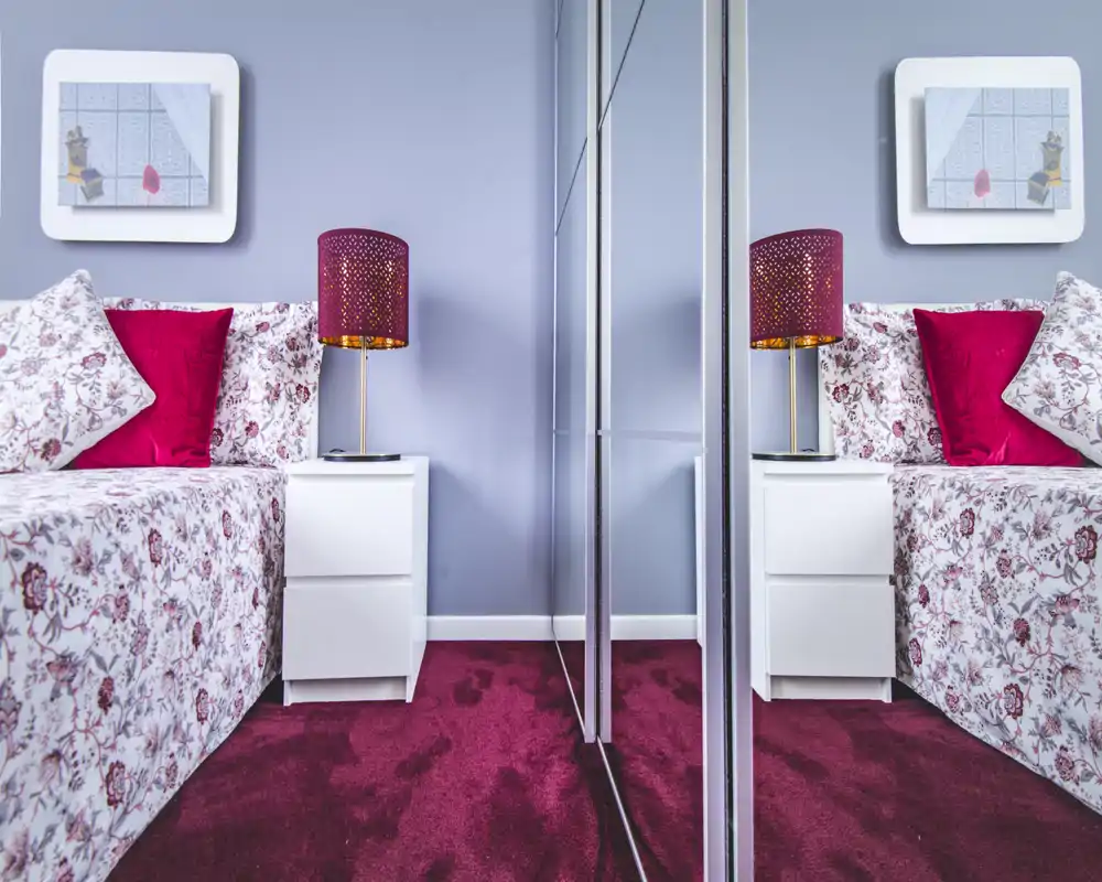 Coin chambre lumineux et élégant avec literie à motifs, moquette rouge douillette et table de chevet avec lampe décorative; penderie miroir apportant profondeur et symétrie.