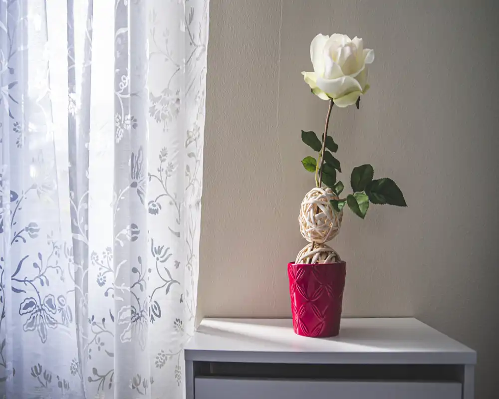 Coin de chambre doucement éclairé avec un voilage à motifs et une unique rose blanche décorative dans un pot rouge posé sur une petite table de chevet blanche. L'image met en valeur une décoration minimaliste et la lumière naturelle.