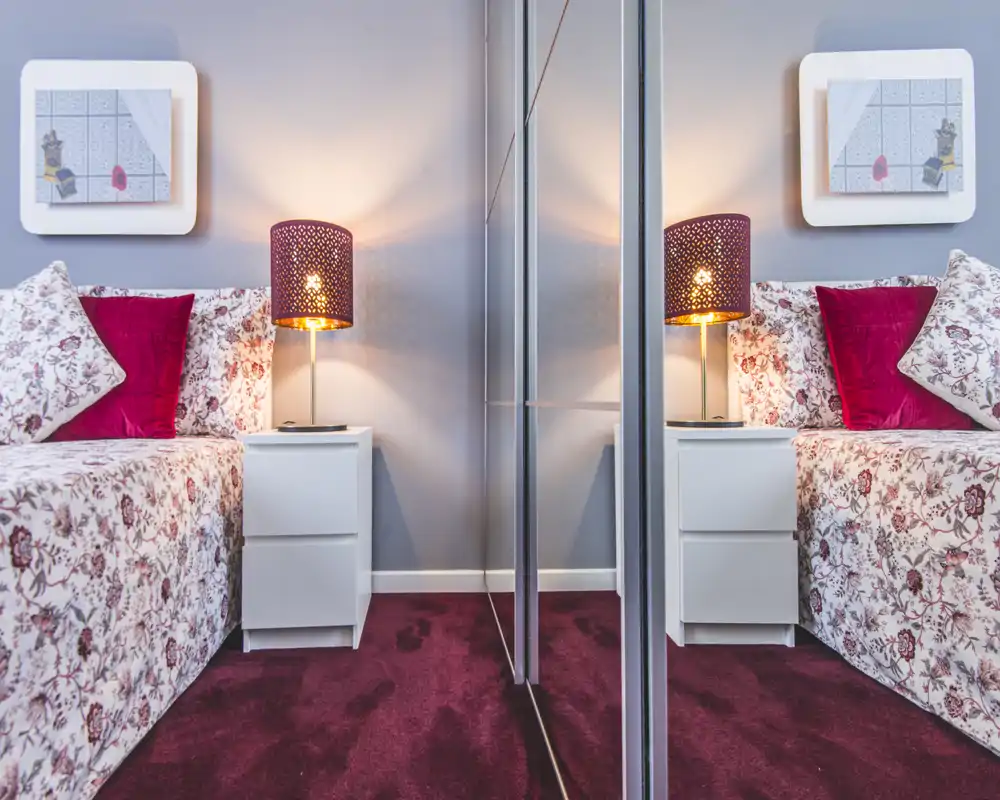 Coin de chambre lumineux et accueillant avec lit à motifs, coussins décoratifs et lampe de chevet ; une armoire miroir apporte profondeur et symétrie.