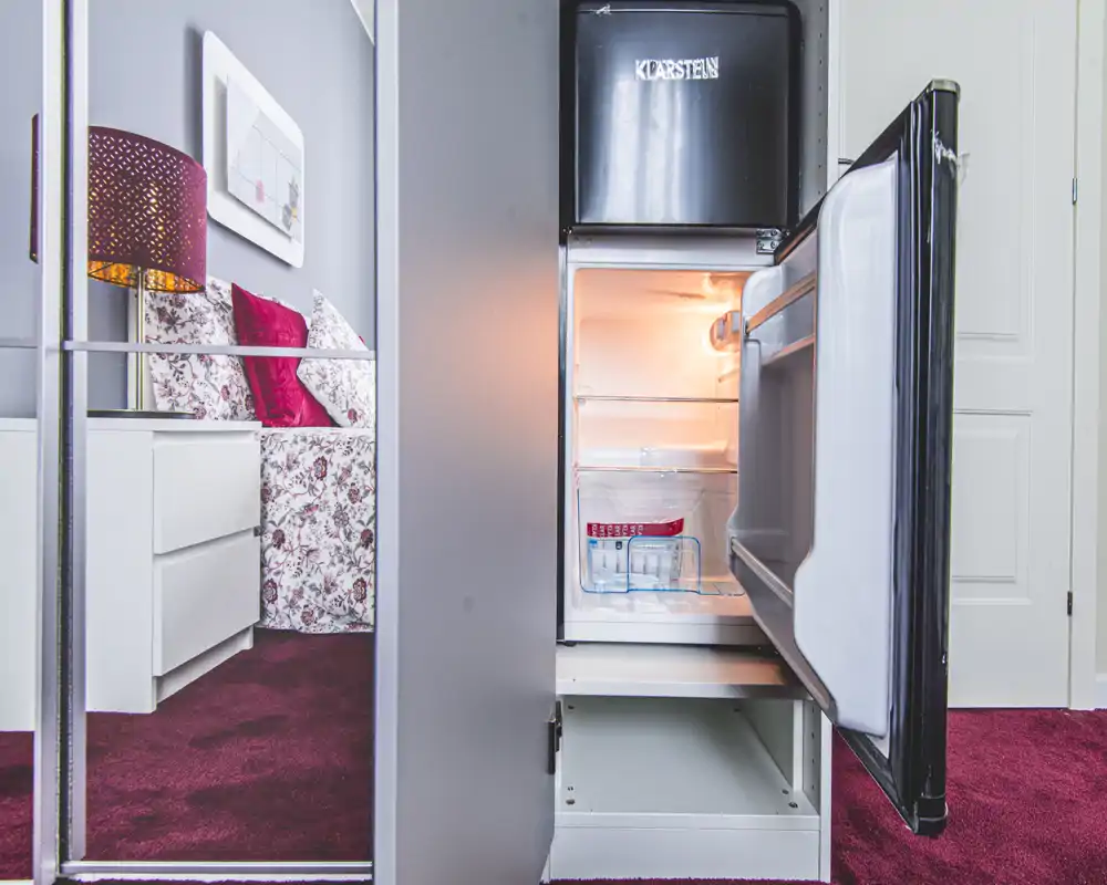 Scène de chambre visible partiellement via la porte miroir de la garde-robe : lit fleuri avec coussins décoratifs et table de chevet avec lampe. Le mini-réfrigérateur ouvert au premier plan suggère des équipements pratiques dans la chambre.