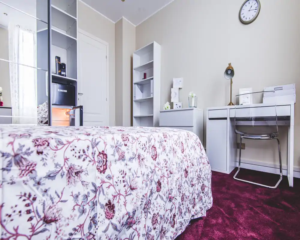 Chambre lumineuse et bien mise en scène avec couvre-lit fleuri, bureau et étagères — idéale pour un séjour confortable.