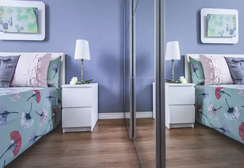 Dormitorio luminoso y con estilo, con edredón estampado, cojines suaves y una mesita blanca con lámpara; el armario con espejo aporta profundidad.