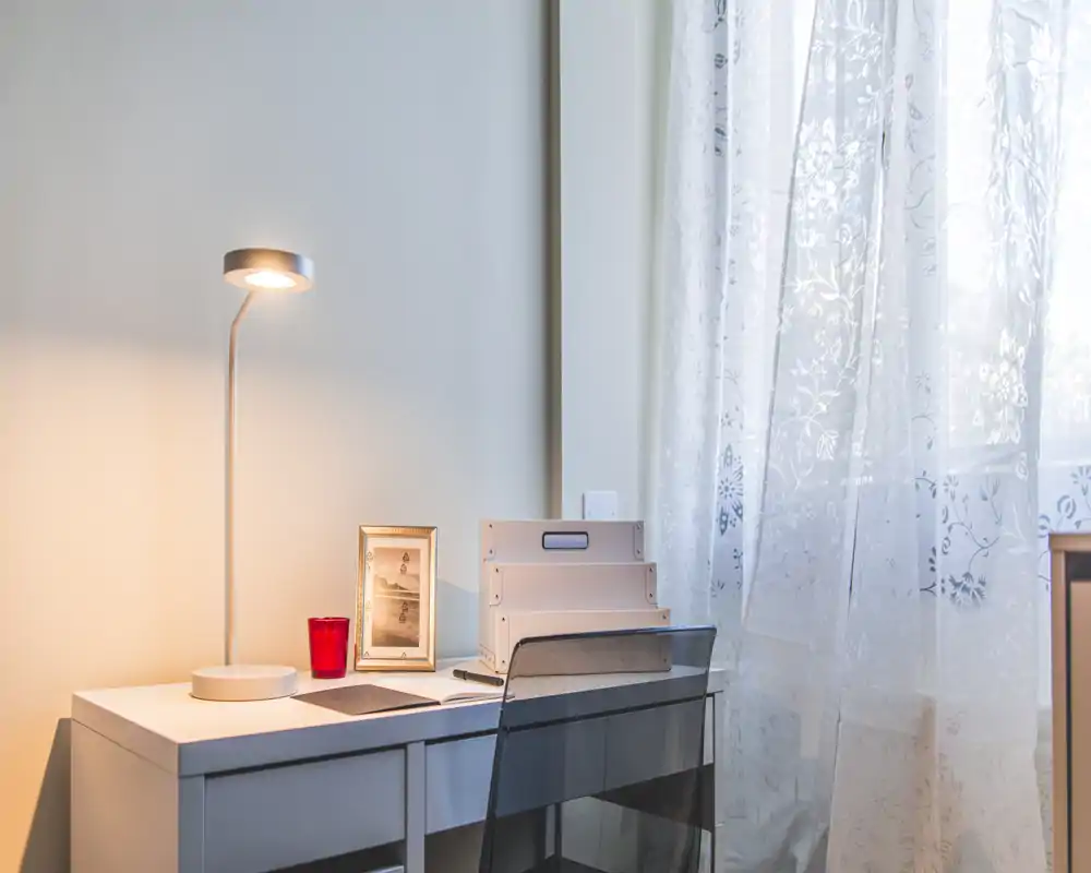 Bright, minimal workspace with a white desk, task lamp, framed photo and stackable organizers beside a sheer curtain—ideal for focused work or study.