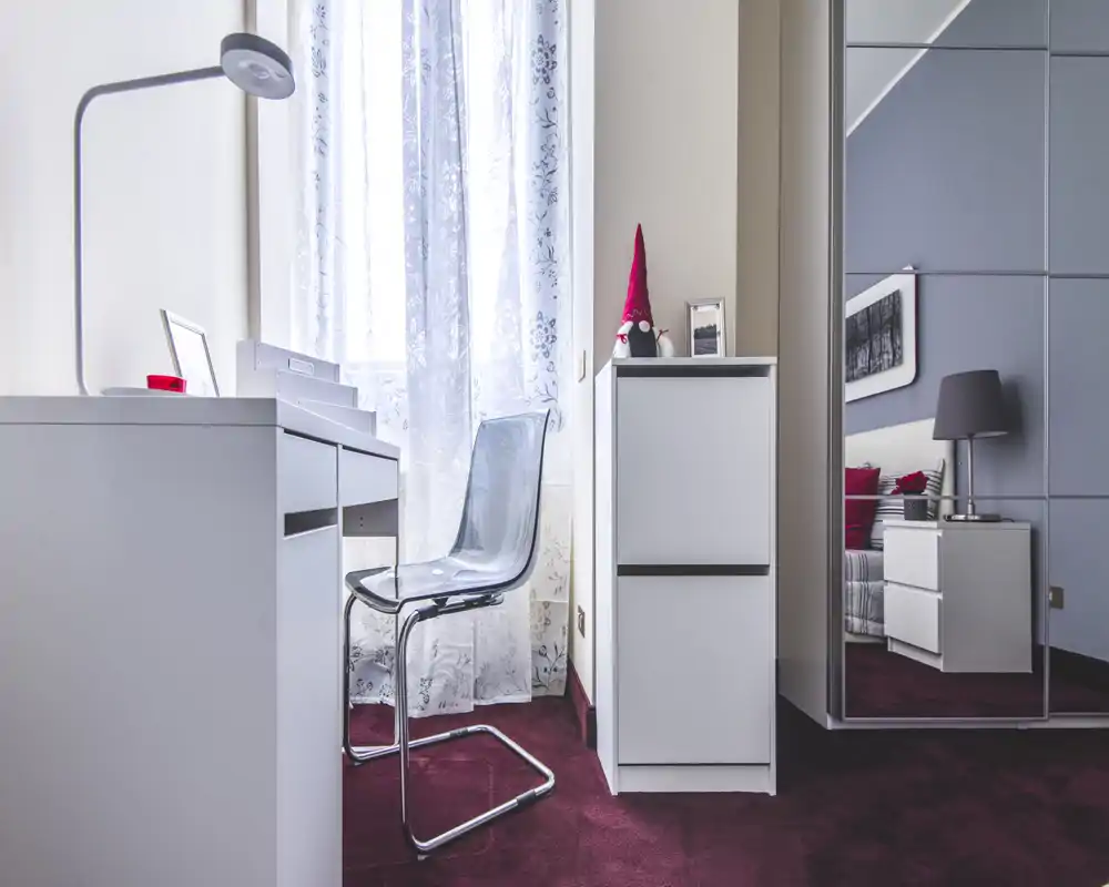 Bright, modern bedroom corner with a white desk, transparent chair and mirrored wardrobe reflecting a bedside table and lamp; decorative textiles and a patterned sheer curtain create a clean, cozy atmosphere.