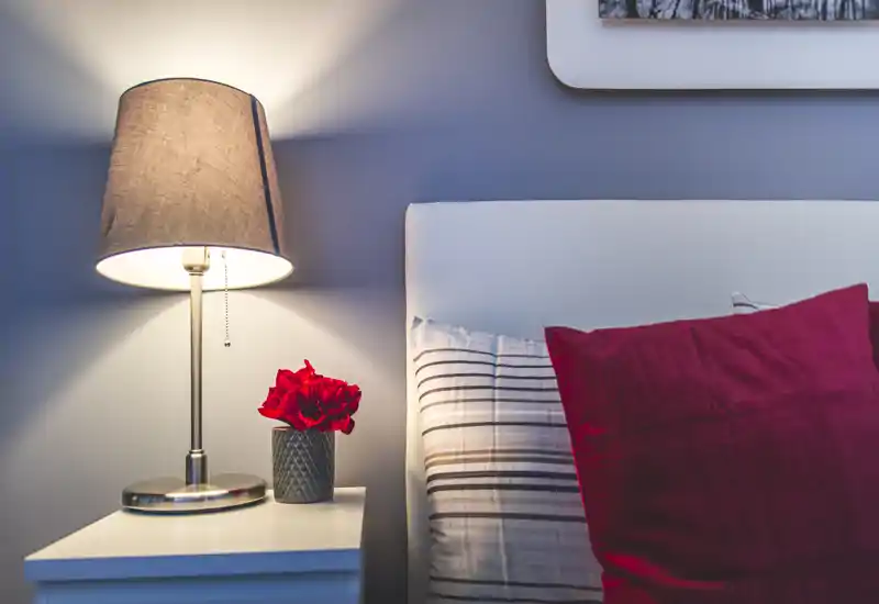 Cozy bedroom detail with a bedside table, lit lamp and a neatly made bed dressed with striped sheets and a red cushion; small decorative flowers add warmth.