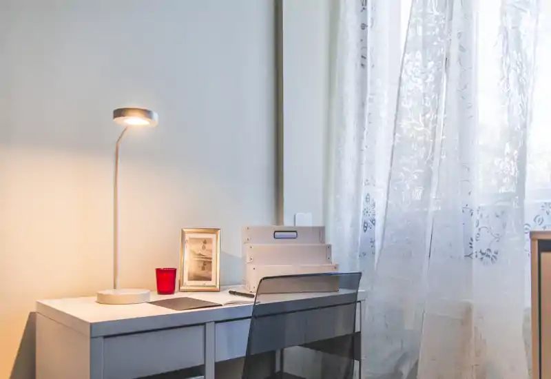 A tidy home office corner with a white desk, task lamp and transparent chair next to a bright window with patterned sheer curtains.