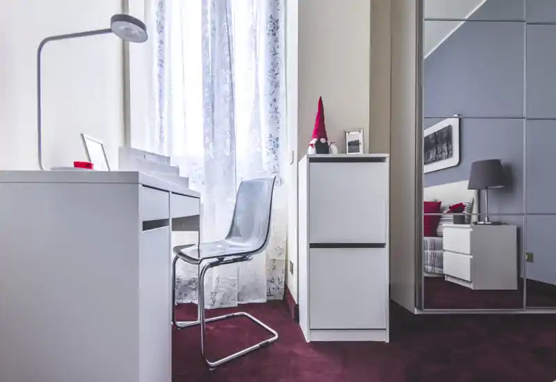 Bright, modern bedroom corner with a white desk, transparent chair and mirrored wardrobe reflecting a bedside table and lamp; decorative textiles and a patterned sheer curtain create a clean, cozy atmosphere.