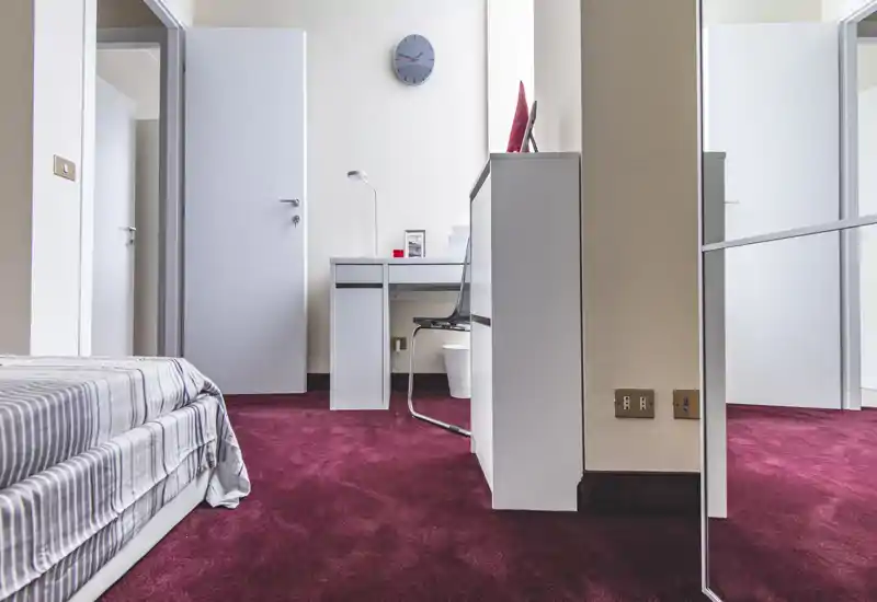 Minimalist bedroom with a neatly made single bed, a compact desk and chair, and a mirrored wardrobe — airy, well lit and quietly styled for a single guest.