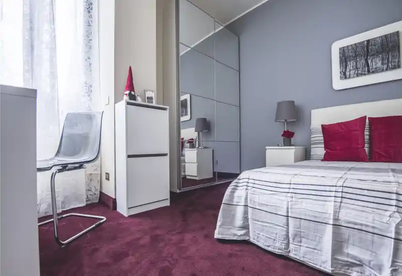 Bright, modern bedroom with a neatly made double bed, red accent cushions, bedside table and a mirrored wardrobe that enlarges the space.