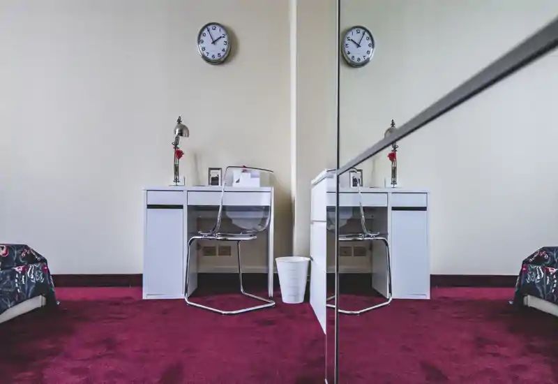 Angolo di camera luminoso con scrivania bianca, sedia trasparente e armadio a specchio che riflette la scena; moquette bordeaux e bordi del letto parzialmente visibili.