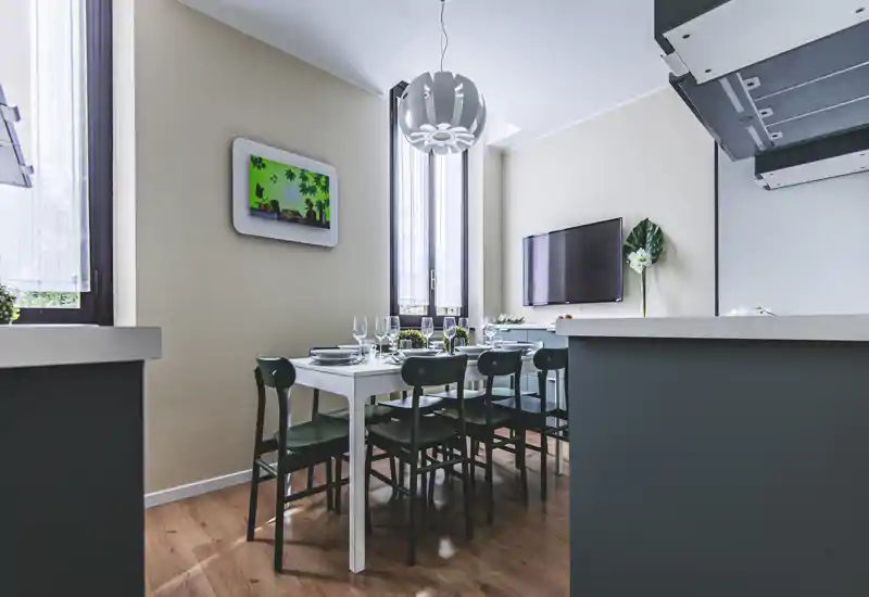 Well-styled dining area with a white table set for guests, six dark-green chairs and tall windows that provide generous natural light. The space integrates with the kitchen island and features warm wooden flooring and modern lighting.
