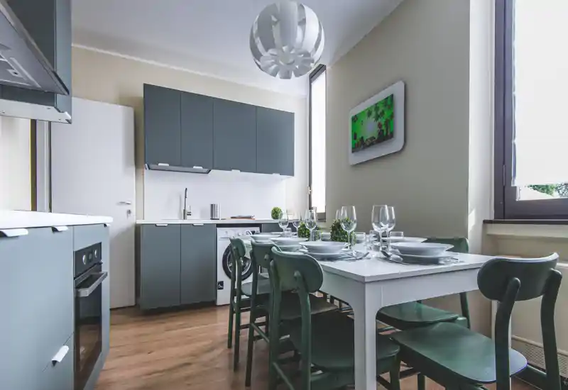 Modern fitted kitchen with a white dining table set for guests, green chairs and coordinated cabinetry; bright, styled and ready for use.