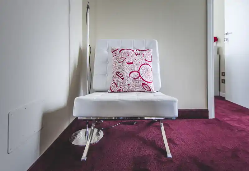 Minimal seating area with a modern white lounge chair, decorative patterned cushion and deep red carpet; compact, stylish corner ideal for relaxing or reading.