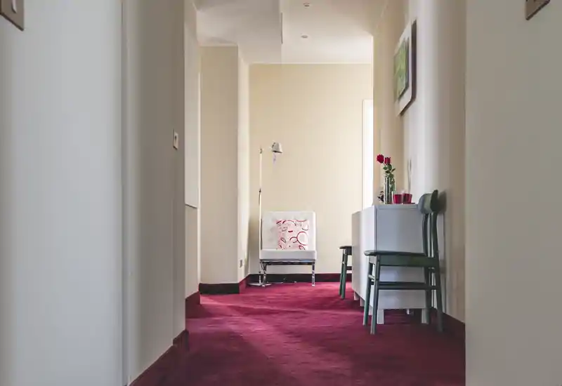 Bright, carpeted entrance corridor with a decorative chair and side storage, styled with a standing lamp and vase — welcoming approach to the apartment.