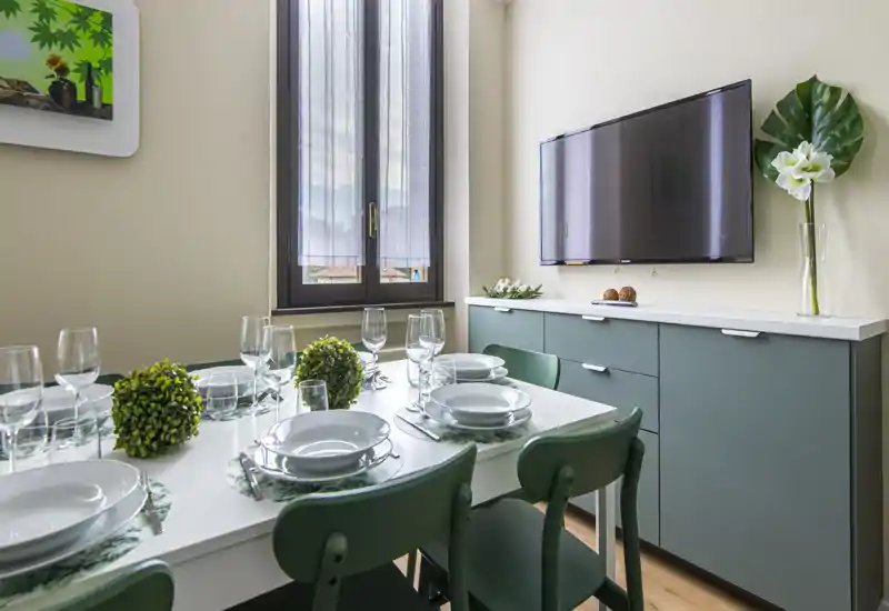 Bright, stylish dining area with a neatly set table for six, green chairs, decorative topiary accents and a sideboard with wall-mounted TV — ideal for shared meals.