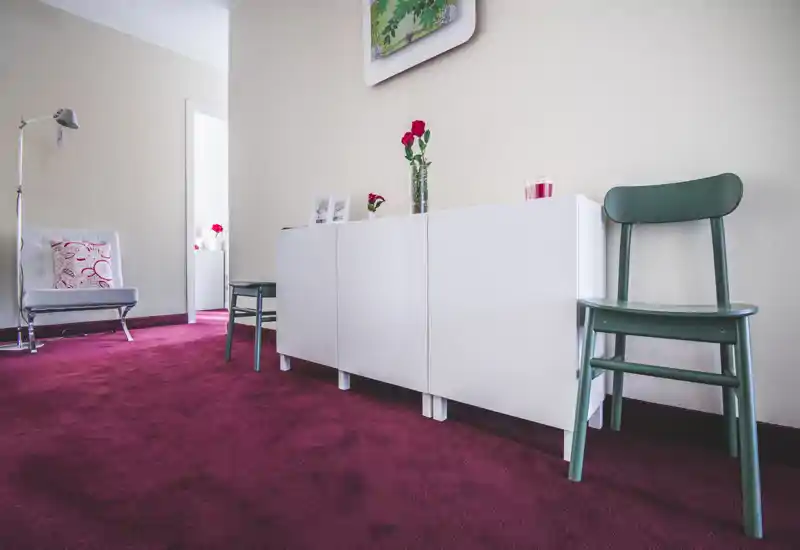 Bright, modern sitting area with a deep-red carpet, minimalist white sideboard, green chairs and decorative flowers — a cozy living space with clean lines and warm accents.