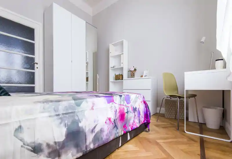 Camera da letto luminosa e moderna con copriletto colorato, mobili bianchi e una scrivania compatta — ideale per una persona o uno studente. Pavimento in parquet e grande armadio con anta specchiata per aumentare la sensazione di spazio.