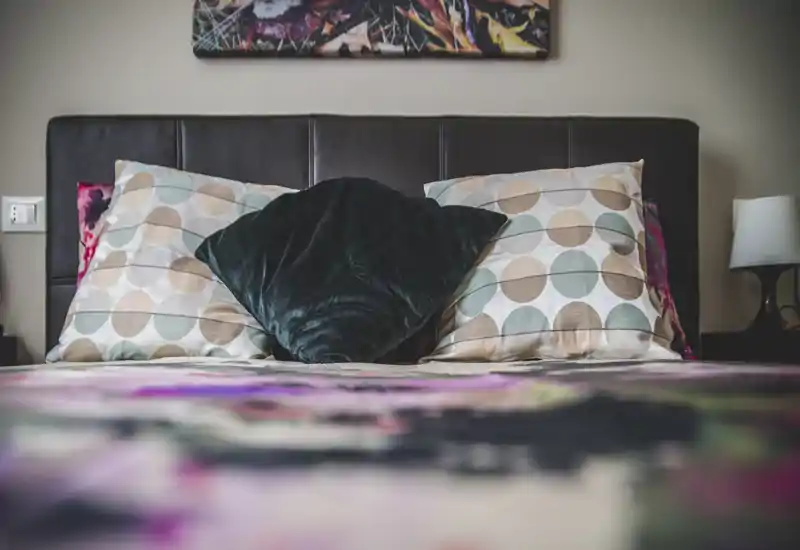 Scatto luminoso e curato della camera da letto incentrato sul letto con cuscini a fantasia, un cuscino in velluto e una testata in pelle — accogliente e ideale per un annuncio.