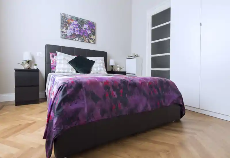 Camera da letto luminosa e curata con un letto matrimoniale rifinito con una coperta viola vivace, cuscini coordinati e comodini con lampade — perfetta per annunci.