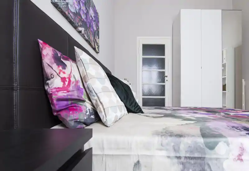 Camera da letto luminosa e ben allestita con un grande letto con biancheria fantasia, cuscini decorativi e un armadio bianco alto sullo sfondo. La composizione mette in risalto comfort e stile moderno, perfetta per annunci di affitto.