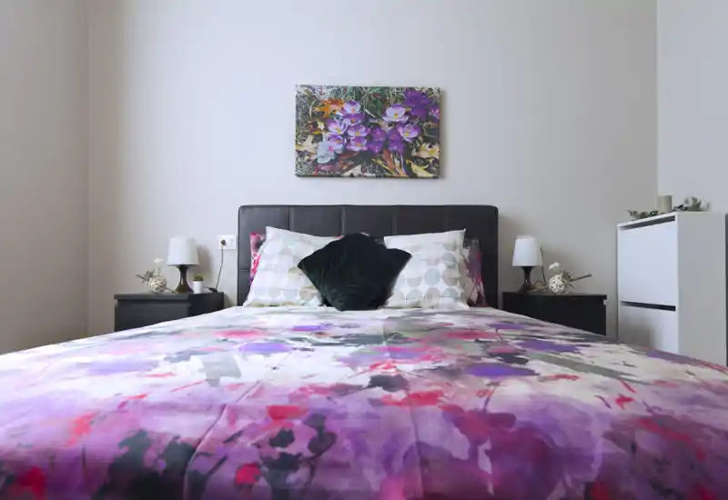Camera da letto luminosa e curata con piumone a fantasia floreale, cuscini decorativi e comodini con lampade — perfetta per annunci che evidenziano uno spazio notte accogliente e ben arredato.