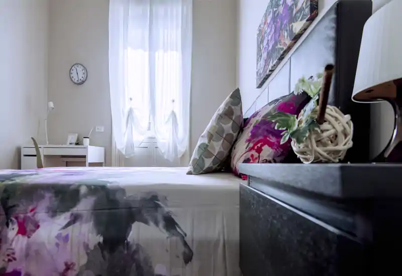 Camera da letto luminosa e curata con copriletto floreale e cuscini decorativi, luce naturale dalla finestra con tende leggere e comodino ordinato con lampada e pianta.
