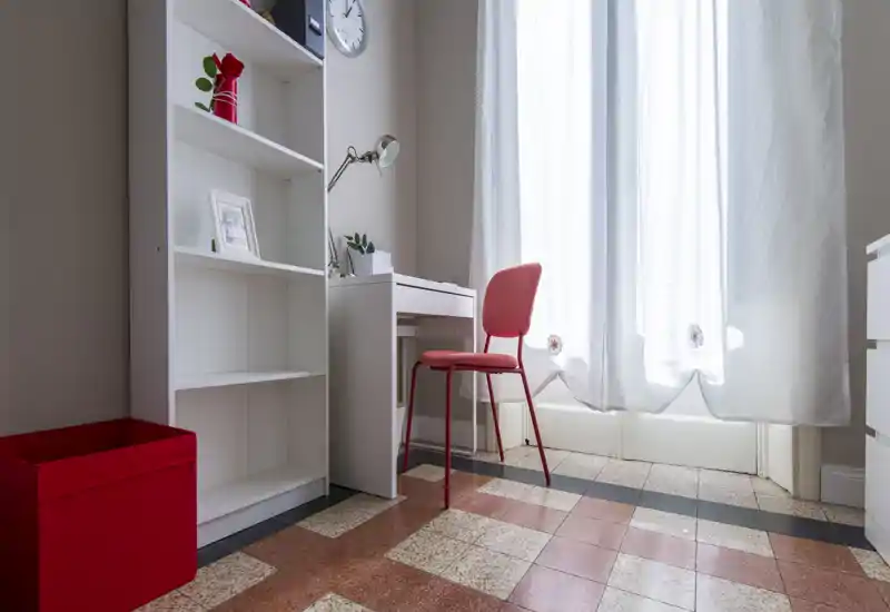 Bright, tidy bedroom corner with a small white desk, red chair and shelving by a sunlit window — ideal as a workspace within the sleeping area.