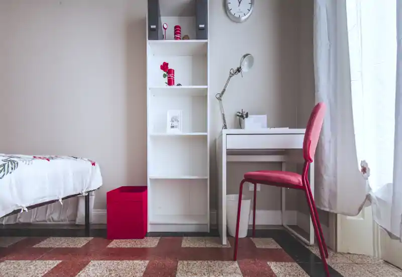 Bright, simply styled bedroom corner with a visible single bed, open shelving and a small white desk with a red chair beside a curtained window.