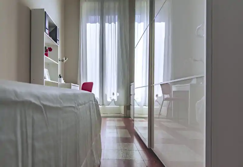 Bright, tidy bedroom with a single bed in the foreground, a desk and shelving by a large window and a glossy wardrobe reflecting light.